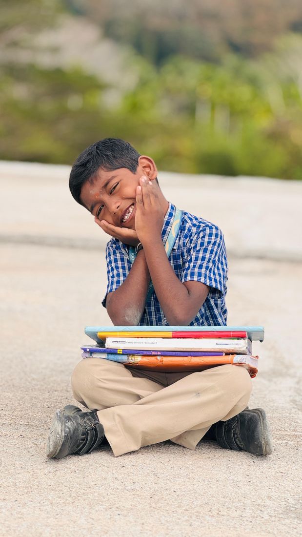 A smiling young student sitting cross-legged with a stack of textbooks on his lap